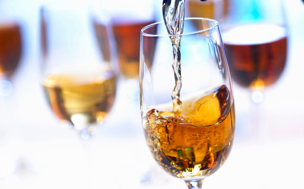 Imagem com 4 taças de vinho de sobremesa e uma em destaque sendo servida. O vinho tem coloração dourada.