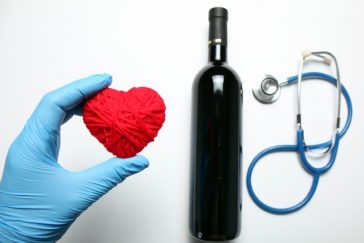 Mão com luva azul segurando coração de pelúcia ao lado de uma garrafa de vinho e estetoscópio representando os benefícios do vinho para saúde