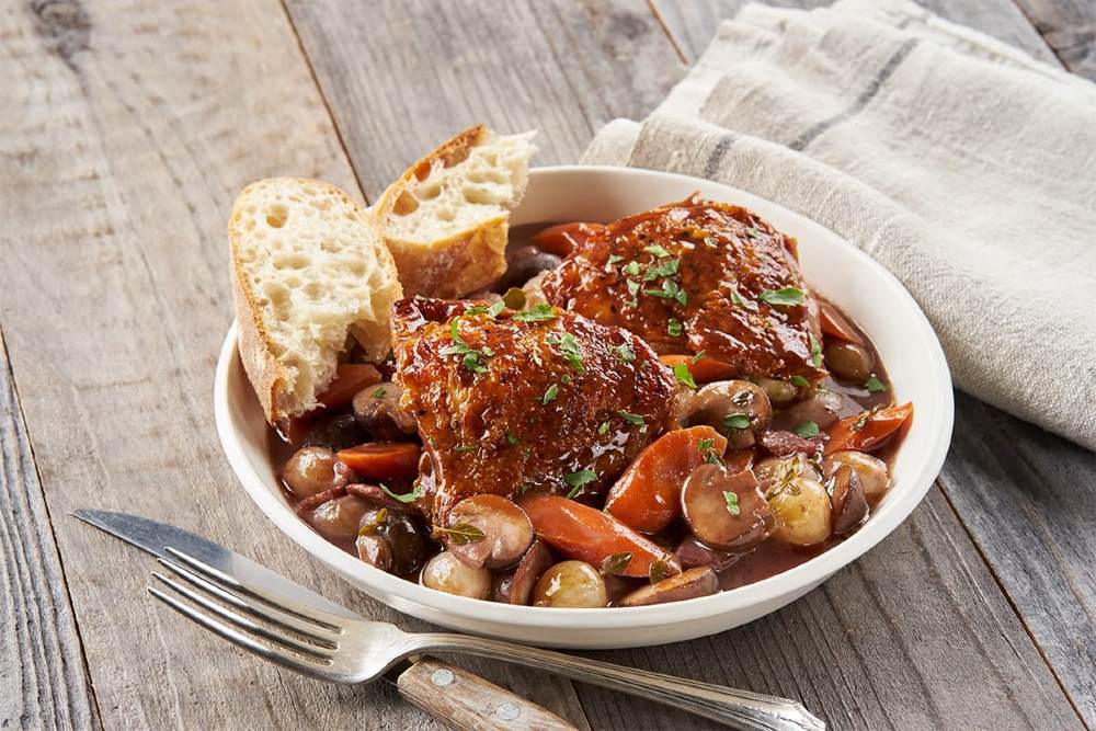 Em uma mesa de madeira clara estão talheres uma toalha branca e um prato branco com a receita de Coq au Vin