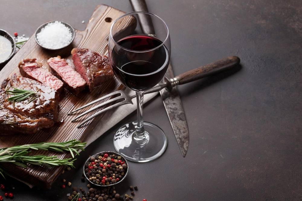 Taça de vinho tinto em cima de uma mesa ao lado de uma tábua de madeira com pedaços de carne mal passadas ilustrando trecho do artigo sobre quais comidas harmonizam com vinhos no inverno