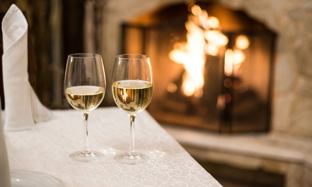 Duas taças com vinho branco em cima de uma mesa com toalha e guardanapos brancos e ao fundo uma lareira acesa ilustrando trecho do artigo que confirma a possibilidade de tomar vinho branco no inverno
