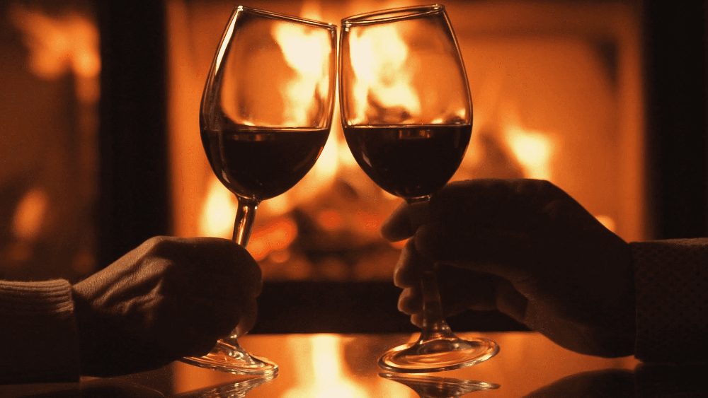 Detalhe de duas pessoas brindando taças de vinho em um cômodo escuro e ao fundo uma lareira acesa ilustrando artigo sobre os melhores vinhos para o inverno